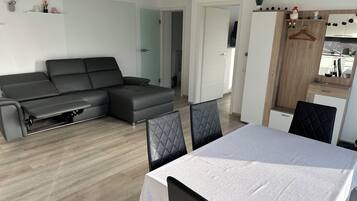 Un salon moderne avec un canapé d’angle, une table à manger avec des chaises et un coin cuisine doté d’un meuble en bois et d’une plante suspendue.