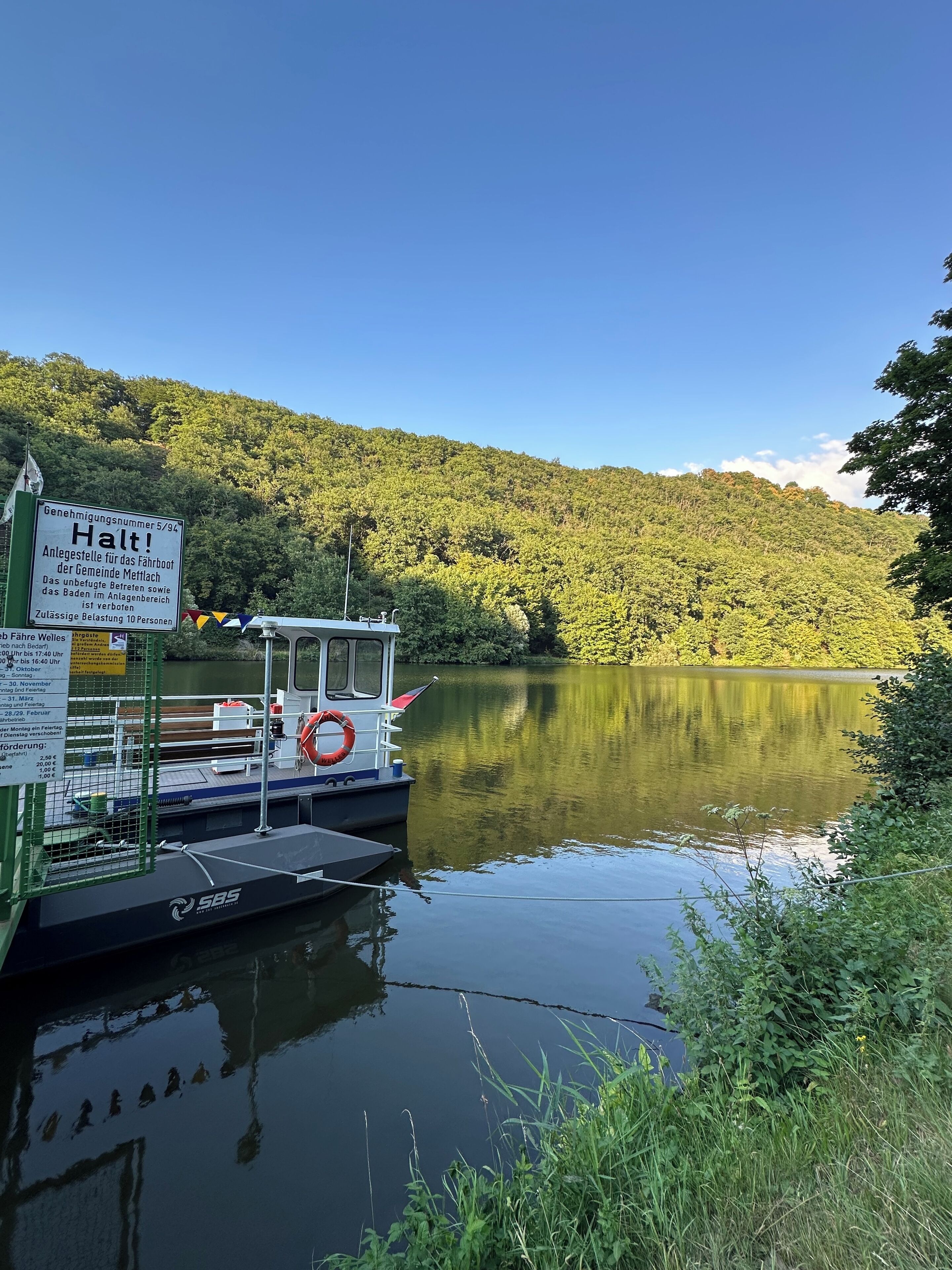 Ein Boot liegt an einem stillen See, im Hintergrund ist ein bewaldeter Hügel zu sehen.