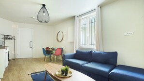 A modern living room with a blue sofa, a wooden coffee table with plants, a dining area with chairs, and a kitchen area with a drying rack. - Apartment Exposition Center (Paris)