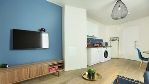 A modern living room with a flat-screen TV mounted on a blue wall, a wooden console table, a round coffee table with a plant, and a kitchen area with a washing machine and a hanging rack. - Apartment Exposition Center (Paris)