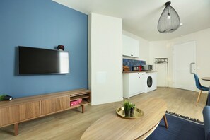 A modern living room with a flat-screen TV mounted on a blue wall, a wooden console table, a round coffee table with a plant, and a kitchen area with a washing machine and a hanging rack. - Apartment Exposition Center (Paris)