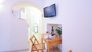 A dining area with a wooden table, chairs, a potted plant, a bottle of wine, and a flat-screen TV mounted on the wall.