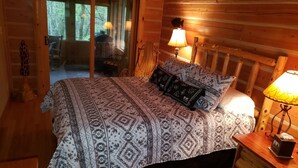 Un dormitorio de cabaña de madera con una cama, dos lámparas de noche y vistas al exterior a través de una puerta de cristal.