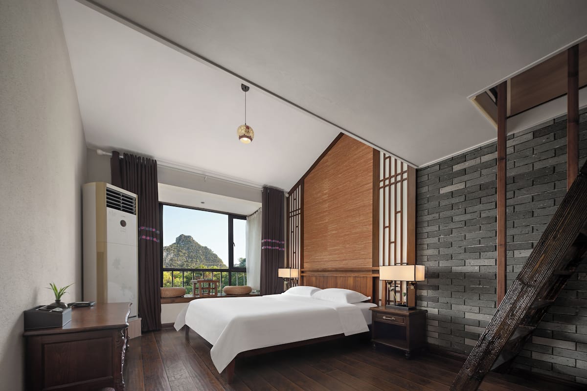A modern hotel room with a large bed, a wooden headboard, a nightstand, a desk, a lamp, and a view of the outdoors.