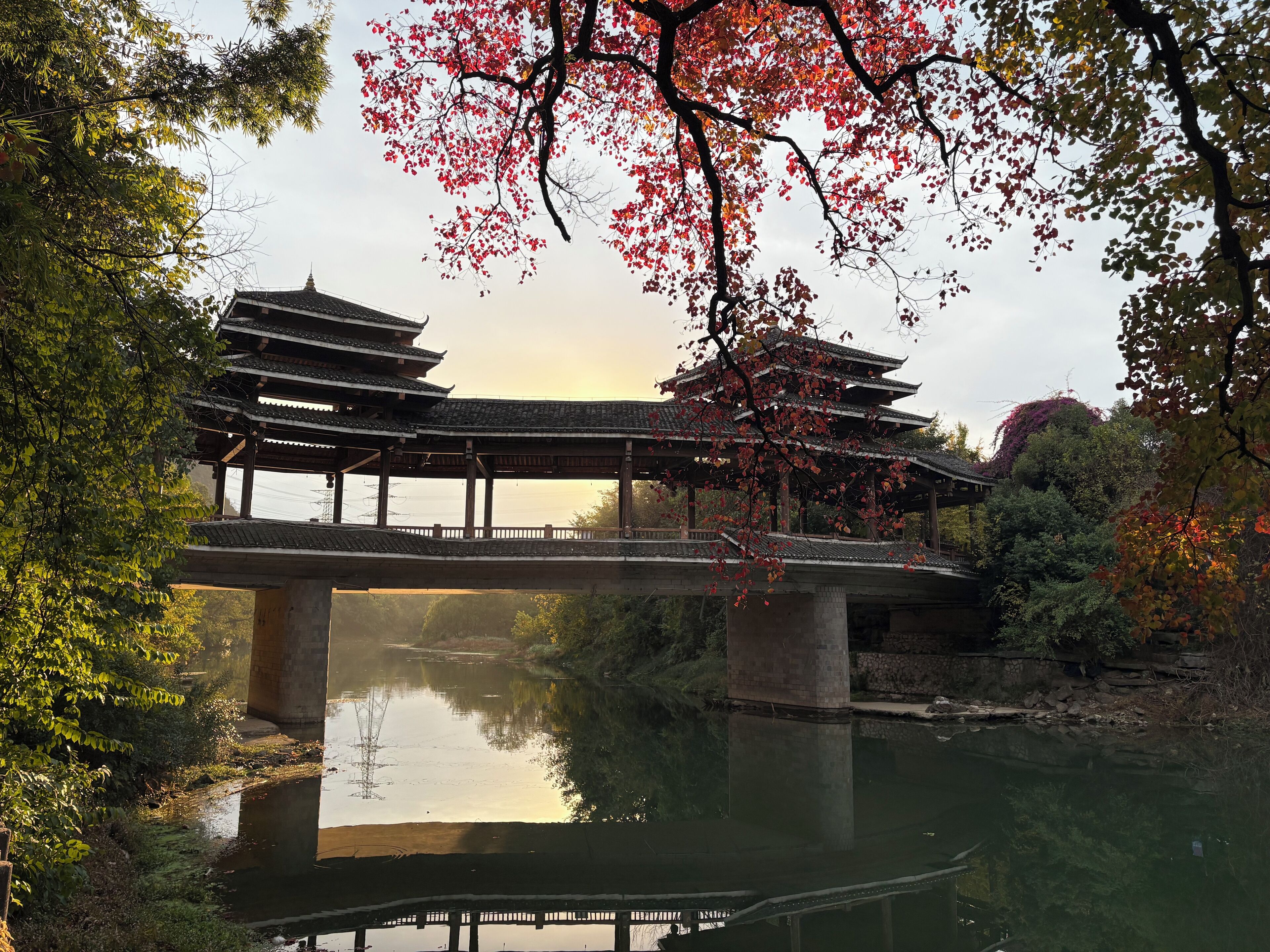 A traditional Japanese bridge over a calm river, surrounded by lush greenery and autumn foliage.