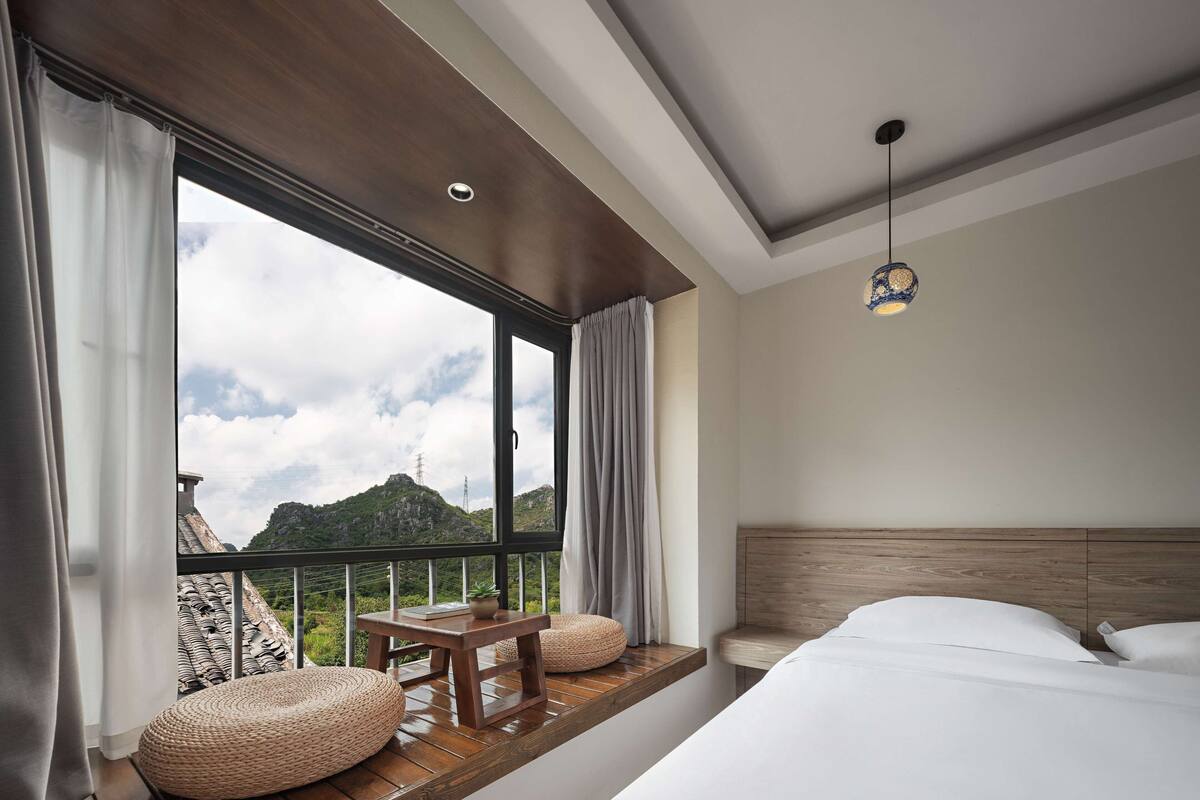 A bedroom with a large window, a wooden headboard, a bedside table, and a hanging light fixture.