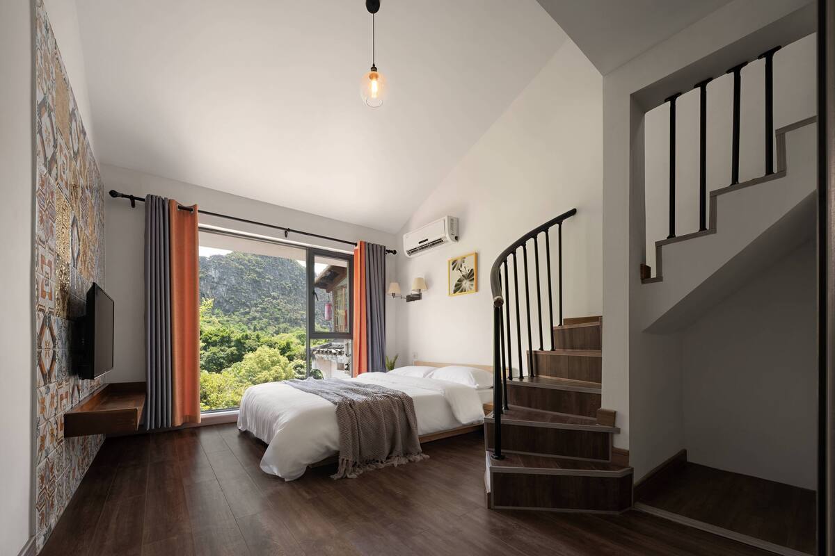 A bedroom with a bed, a TV, a window with curtains, a staircase, and a view of greenery.