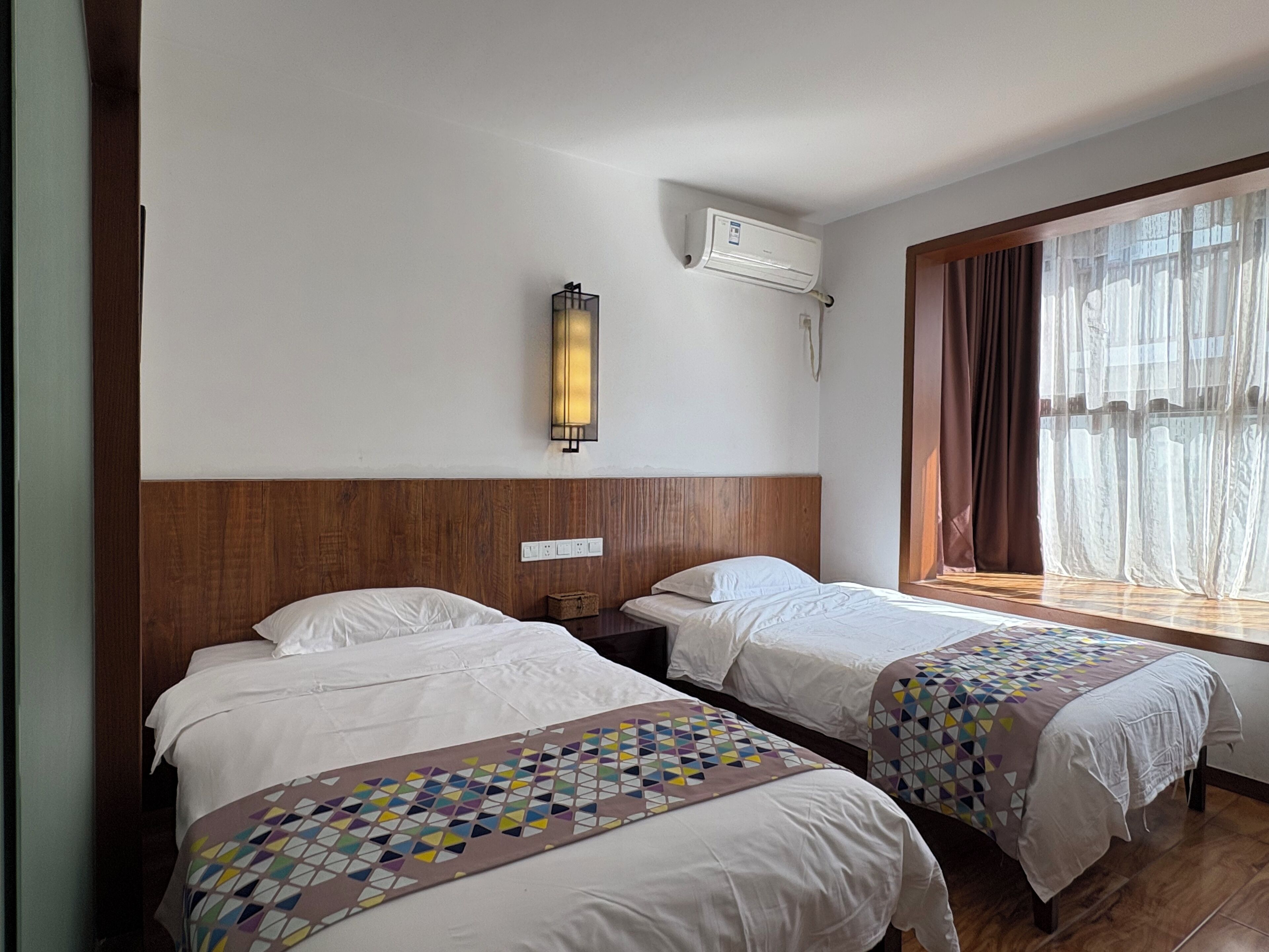 A hotel room with two beds, a wooden headboard, a wall-mounted air conditioner, and a window with curtains.