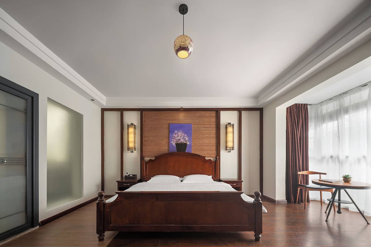 A hotel room with a large wooden bed, two wall-mounted lamps, a ceiling light, a painting on the wall, and a small table with a chair.