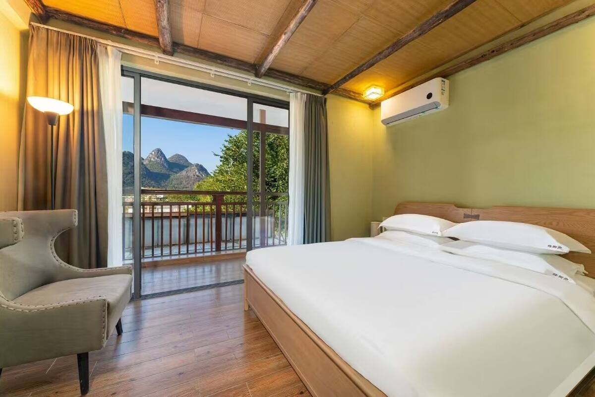 A hotel room with a bed, a chair, a balcony with a view of mountains, and a wooden ceiling.
