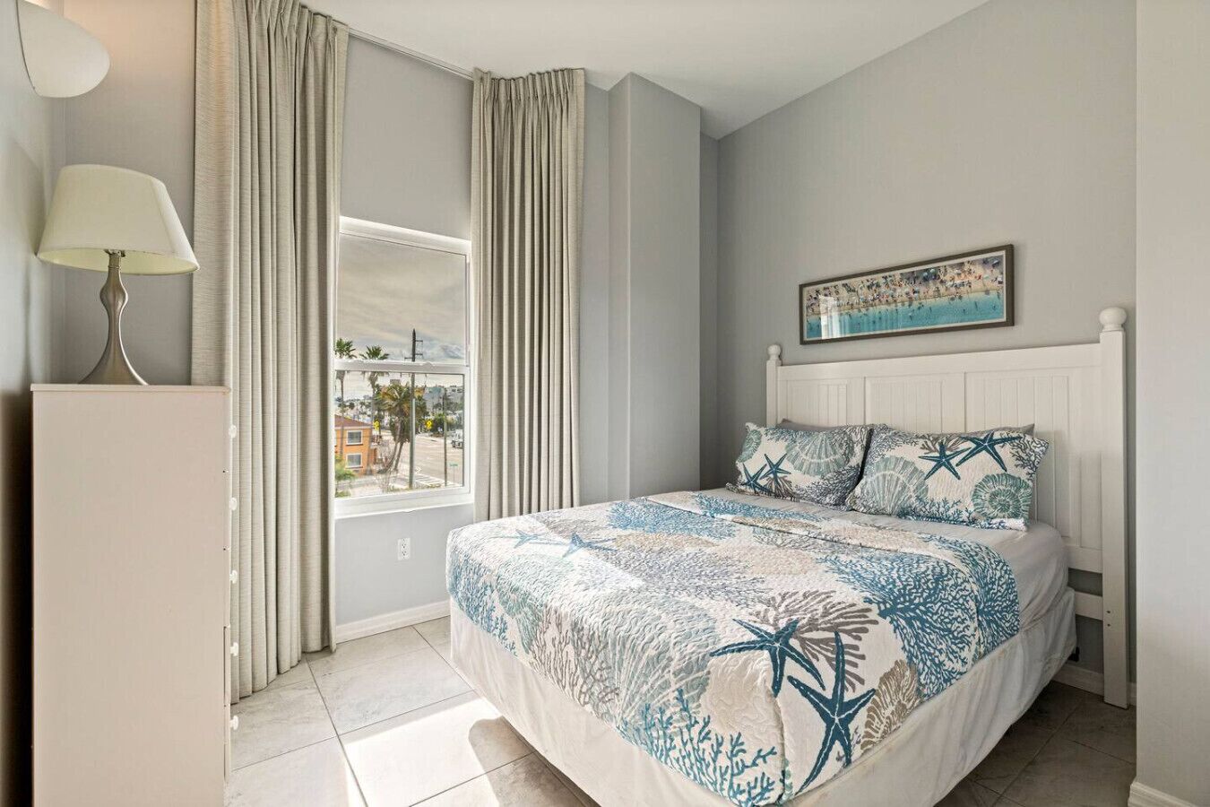 A bedroom with a bed, a window, curtains, a lamp, and a painting.