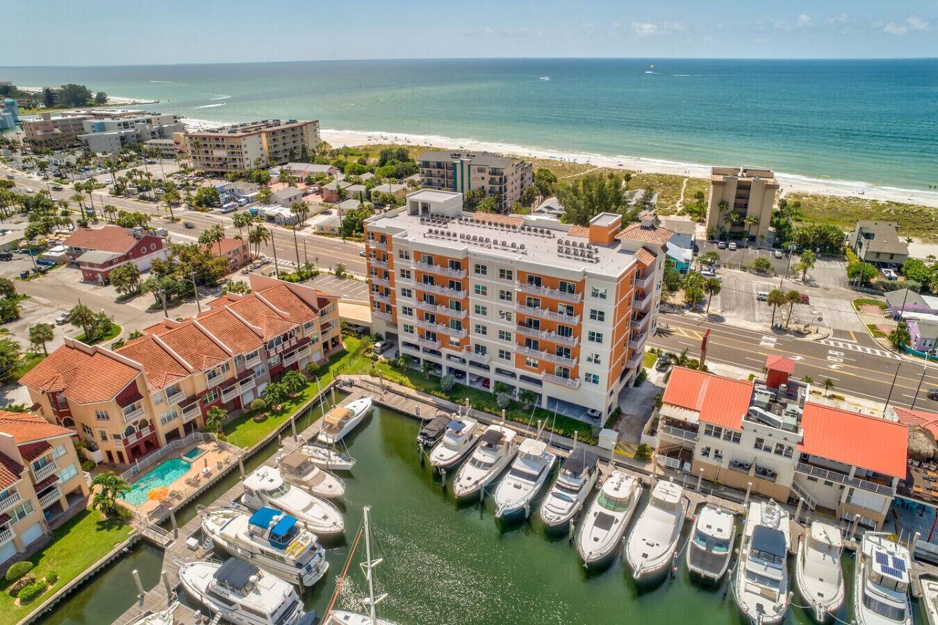 A marina with multiple boats docked, a central building with balconies, and a beach in the background.