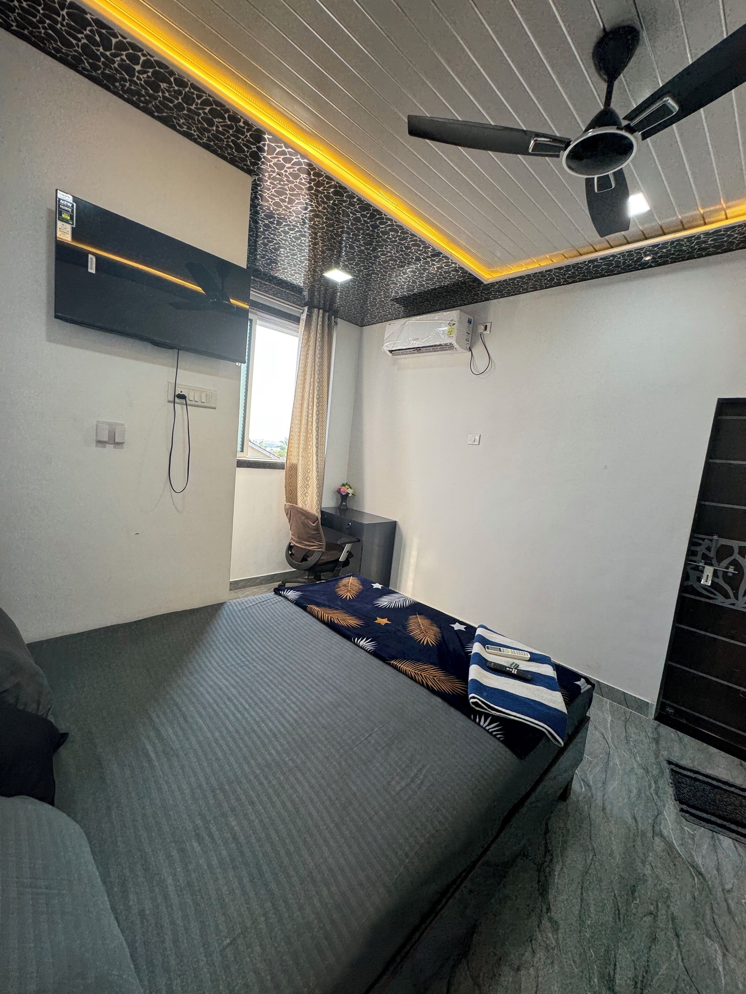 Egy hálószoba, amelyben található egy ágy, egy mennyezeti ventilátor, egy légkondicionáló és egy függönnyel ellátott ablak.