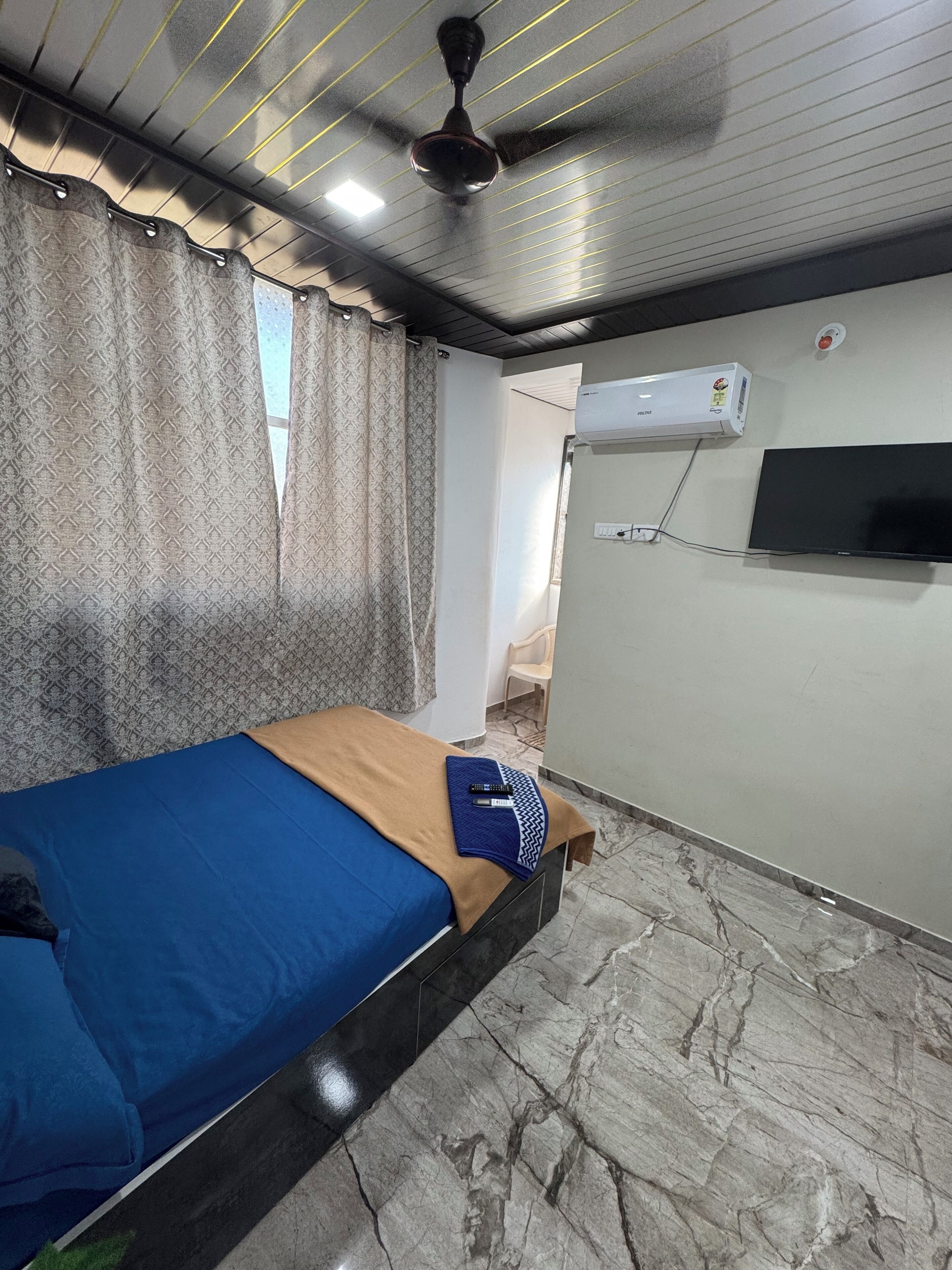 A room with a bed, a ceiling fan, an air conditioning unit, and a TV mounted on the wall.