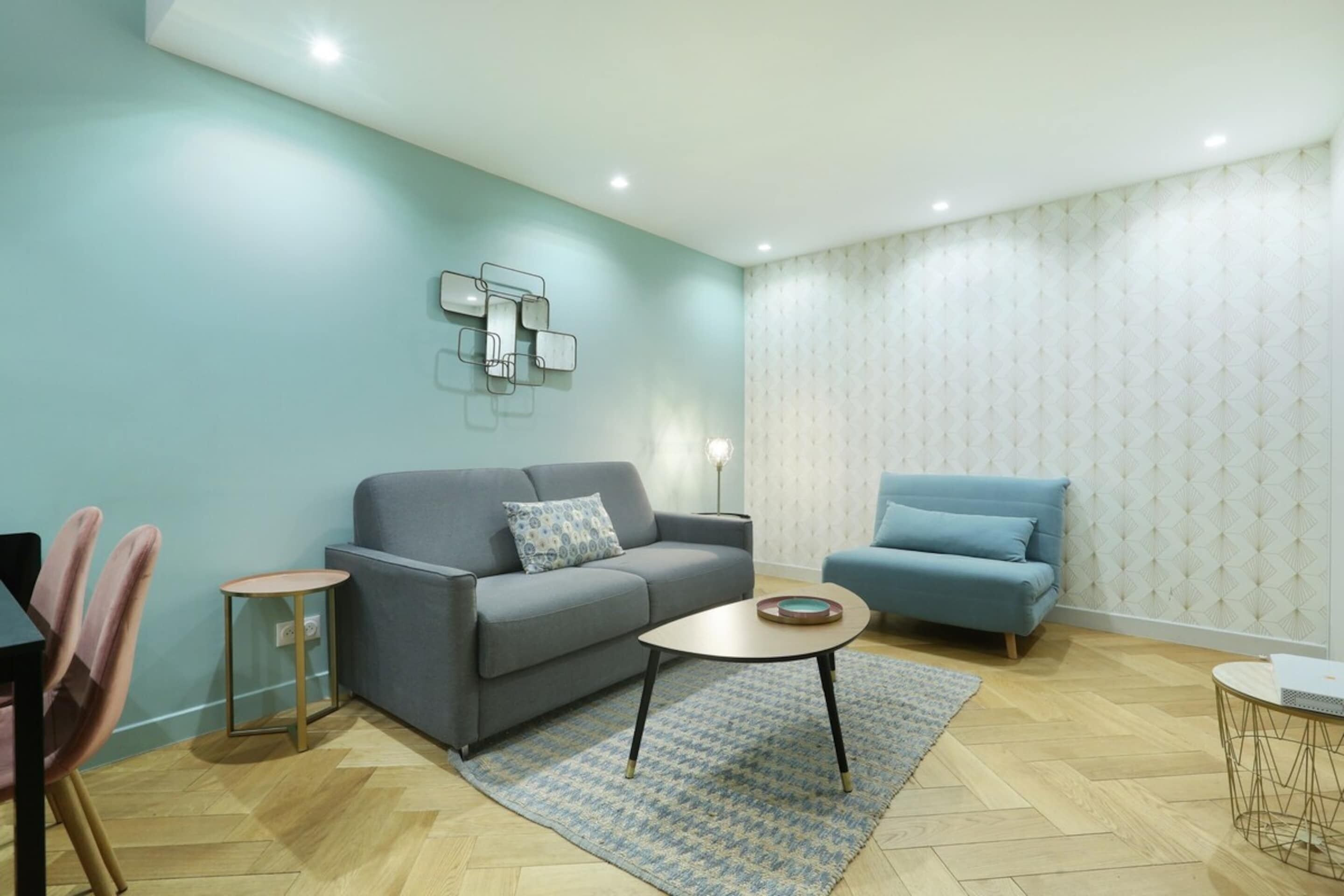 Un salon moderne avec un canapé gris, un fauteuil bleu, une petite table ronde et un tapis à motifs.