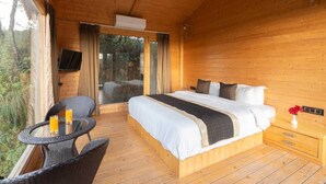 A wooden cabin bedroom with a bed, a small table with chairs, and a glass door leading to an outdoor area. - Nirvana Dhar Nature Resort (Rajgarh)