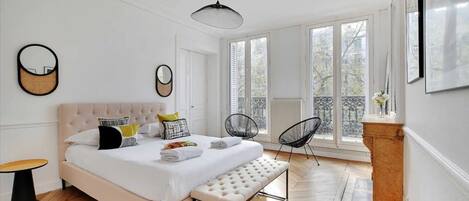 A bedroom with a large bed, a bench, two chairs, a radiator, and large windows.