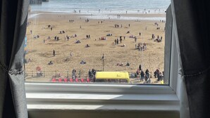 Une vue sur la plage, prise depuis une fenêtre avec des rideaux, montrant une plage de sable, des personnes et un véhicule jaune.