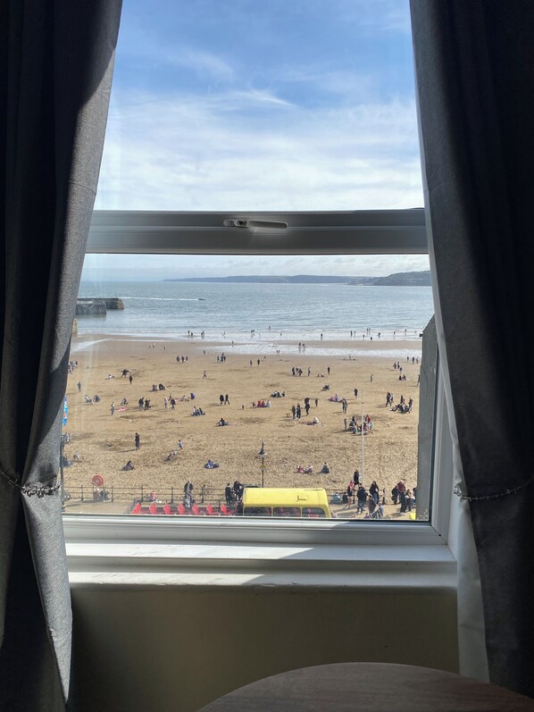A beach view from a window with curtains, featuring a sandy beach, people, and a yellow vehicle.