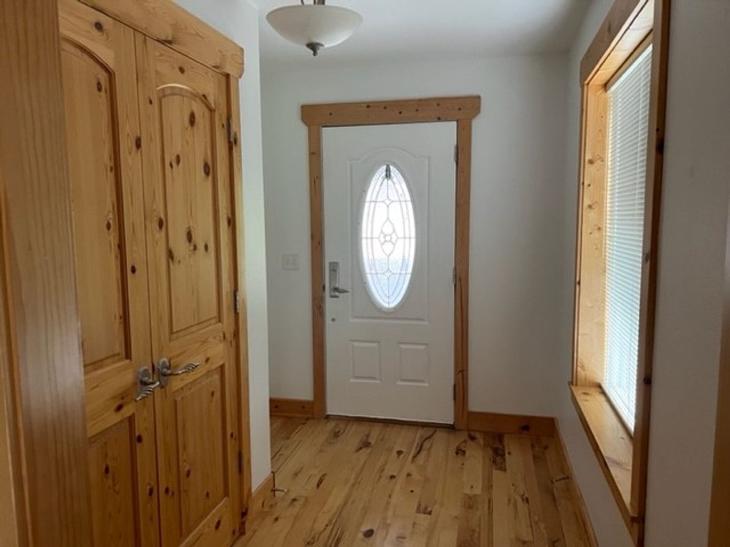 A hallway with wooden flooring, a white door with a glass panel, and a window with blinds.