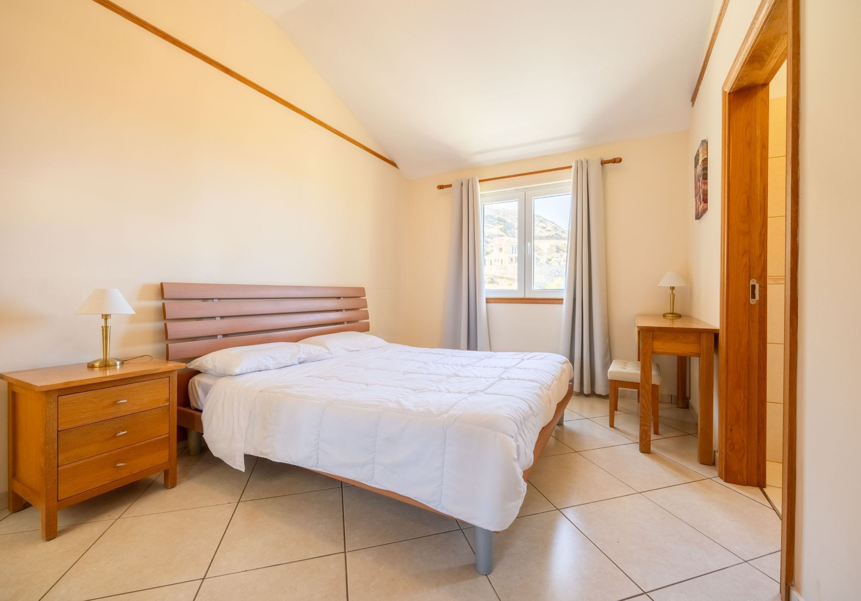 A bedroom with a bed, wooden nightstands, a small table, and a window with curtains.
