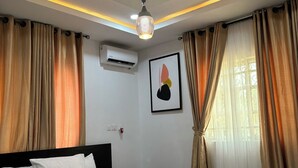 Une chambre à coucher comprenant un lit, des rideaux, un appareil de climatisation, un luminaire au plafond et une œuvre d’art accrochée au mur.