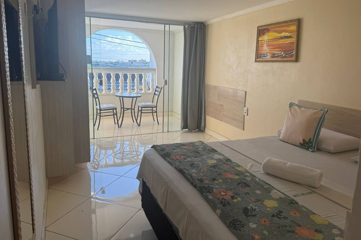 A hotel room with a bed, a painting, a balcony with chairs, and a view of the city.