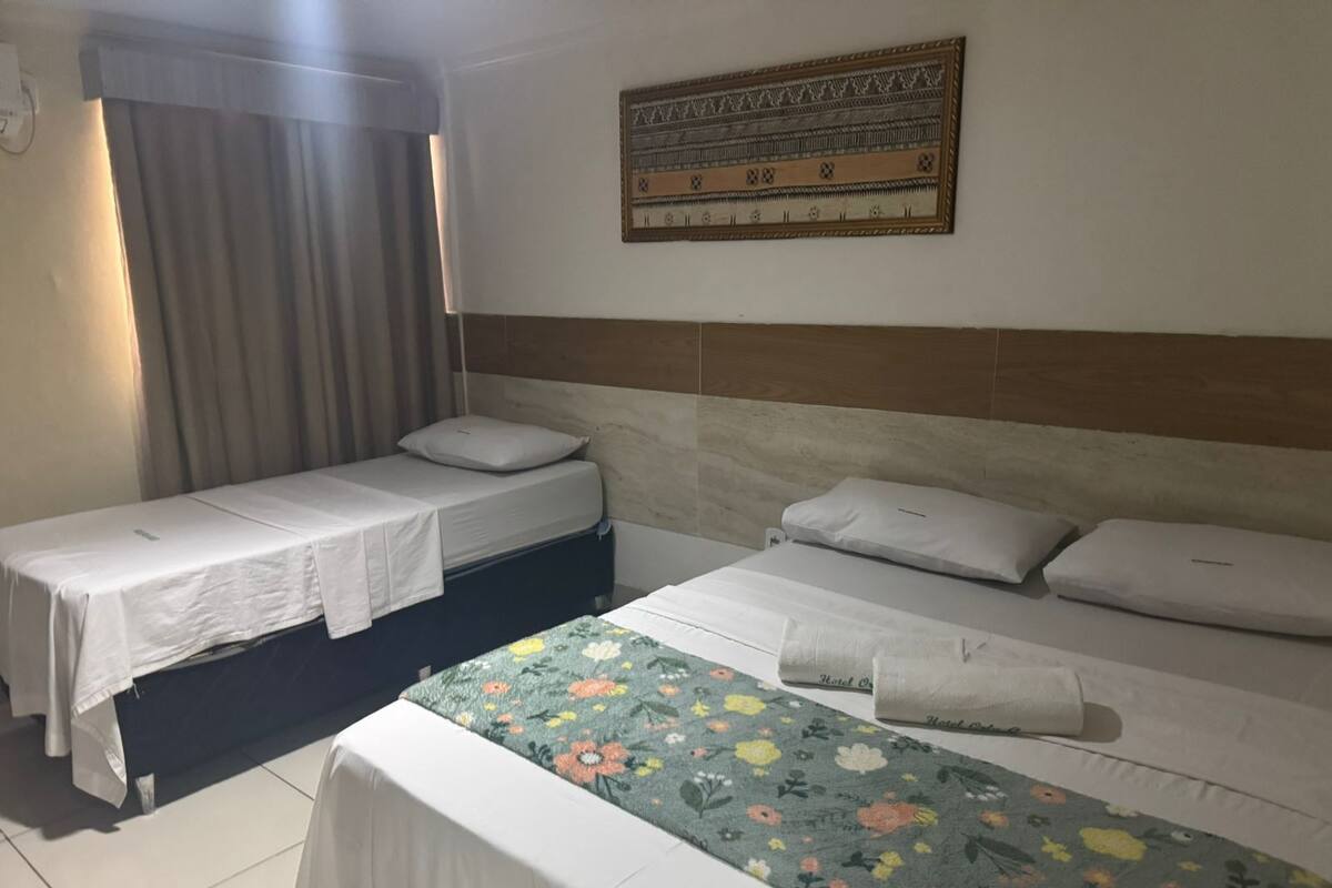 A hotel room with two single beds, a large bed with a floral bedspread, a headboard, a wall-mounted artwork, and a window with curtains.