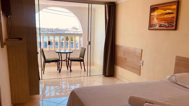 A hotel room with a bed, a small table with chairs, a painting on the wall, and a view of a balcony with a railing. - Hotel orla Guest House (Petrolina)