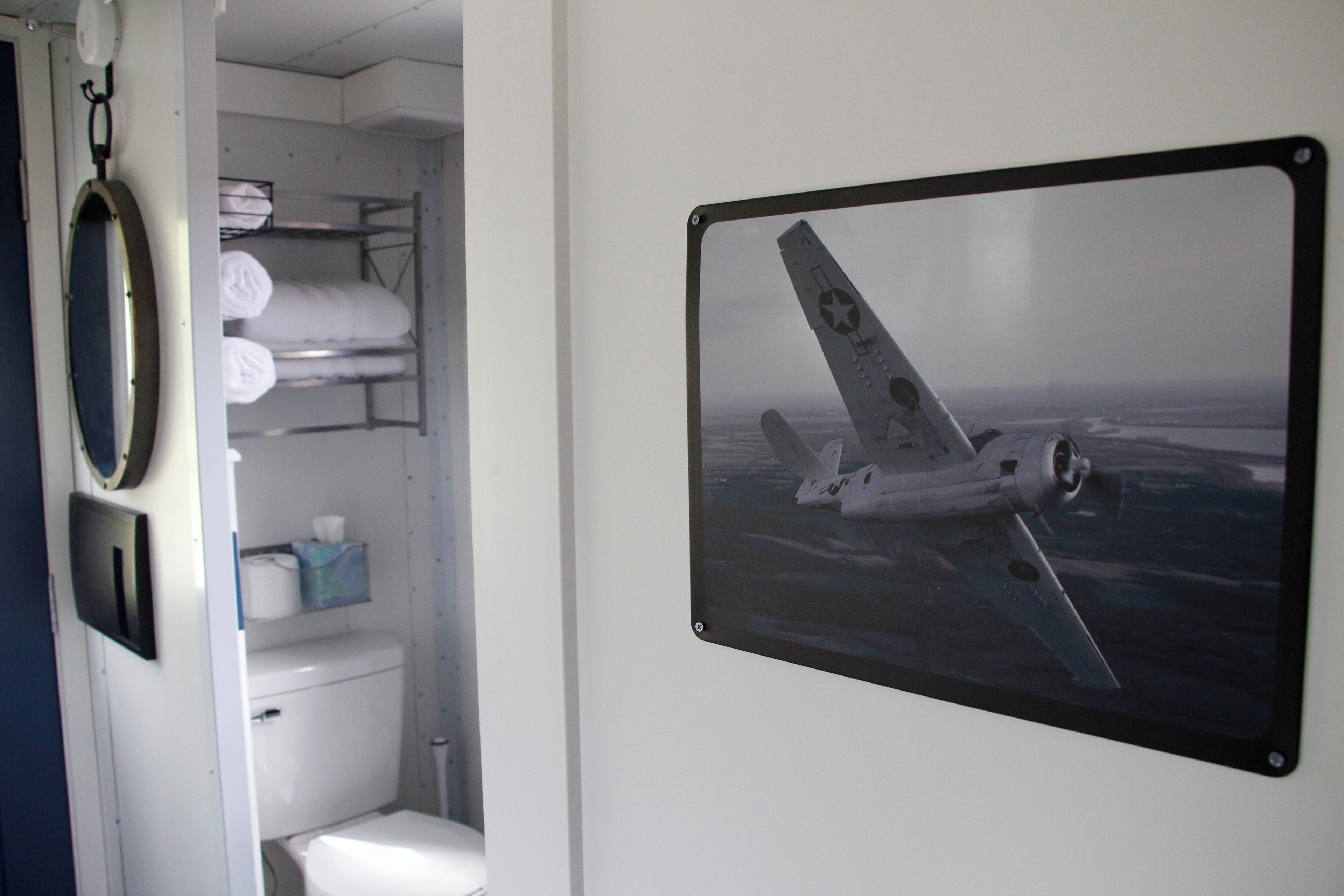A bathroom with a toilet, shelves with towels, and a framed picture of an airplane.