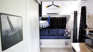 A compact living space with a sofa bed, a hanging hammock, a mounted air conditioner, and a kitchen area with a wooden countertop.