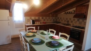 A dining area with a table set for four, surrounded by white chairs with green cushions. A kitchen with wooden cabinets and a tiled backsplash is visible in the background. - DALI HOLIDAY HOME 5 minutes from the sea and the cetacean sanctuary (Imperia)