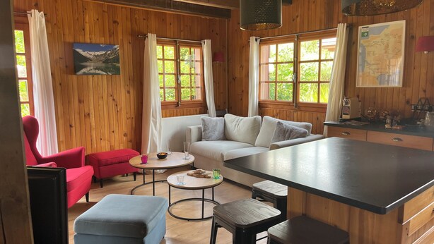 A cozy cabin interior with a wooden interior, a sofa, a red chair, a blue ottoman, a kitchen area, and a map on the wall.