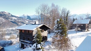 A wooden cabin with a sloped roof, surrounded by snow-covered trees and mountains in the background.