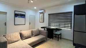 A modern living room with a sectional sofa, a dining table with chairs, and a built-in kitchen area with a refrigerator. - ORYZ APARTMENT (Abuja)
