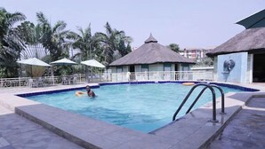 A swimming pool with a thatched-roof structure, palm trees, and umbrellas. - ORYZ APARTMENT (Abuja)