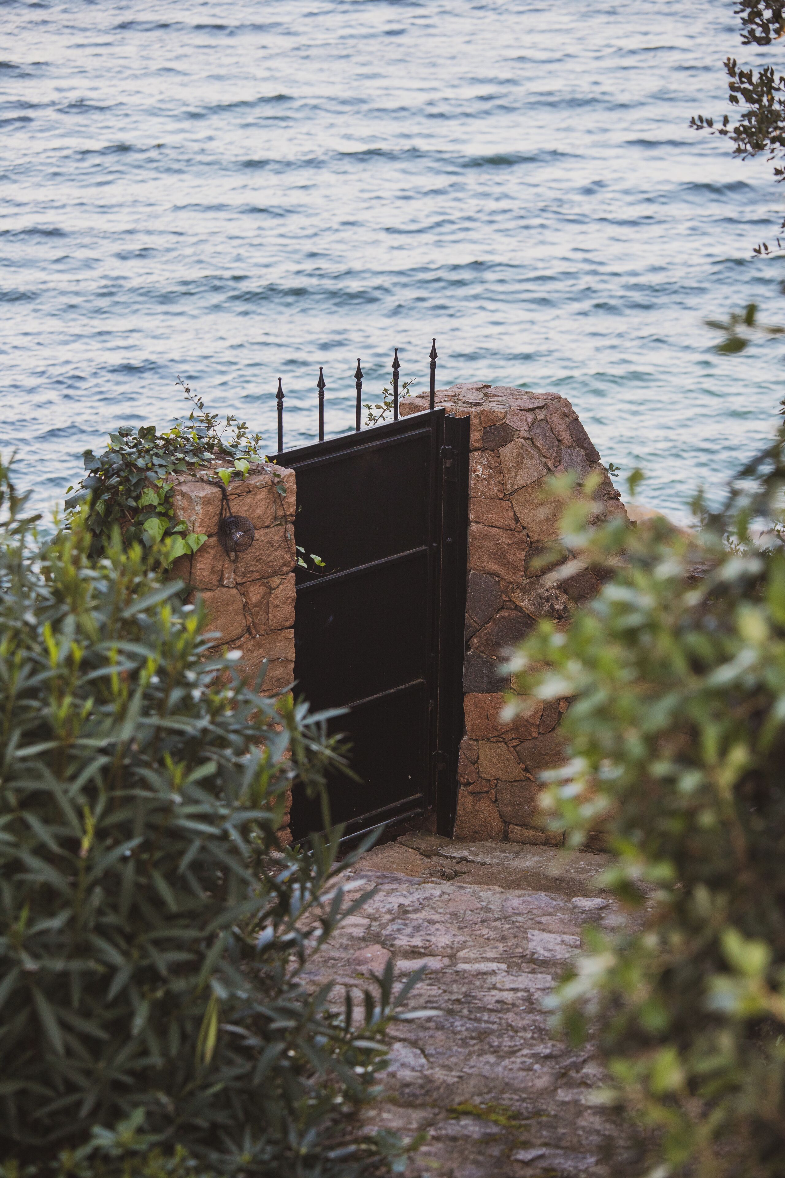 A stone structure with a black metal gate, surrounded by greenery and overlooking a body of water.