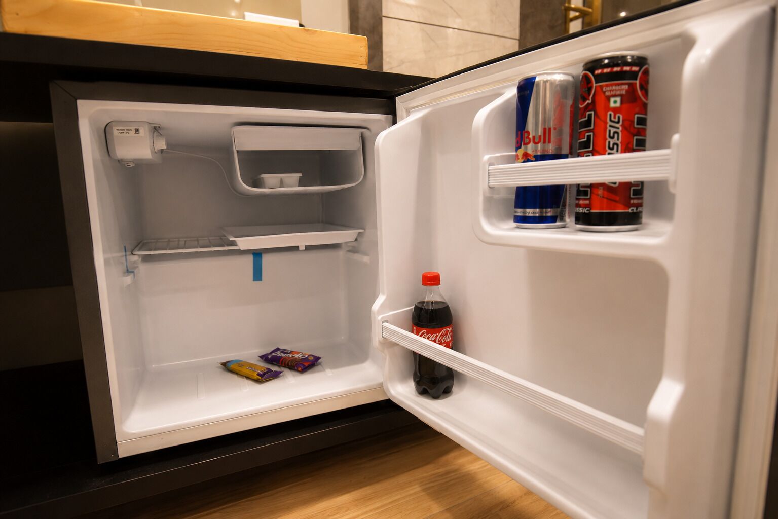 An open refrigerator with a bottle of Coca-Cola, two Red Bull cans, and a Cadbury chocolate bar.