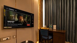 Hotellihuone, jossa on seinälle asennettu taulutelevisio, josta näkyy elokuva, työpöytä tuolilla, pieni pöytä, jossa on puhelin ja kaukosäädin, sekä verhoilla peitetty ikkuna.