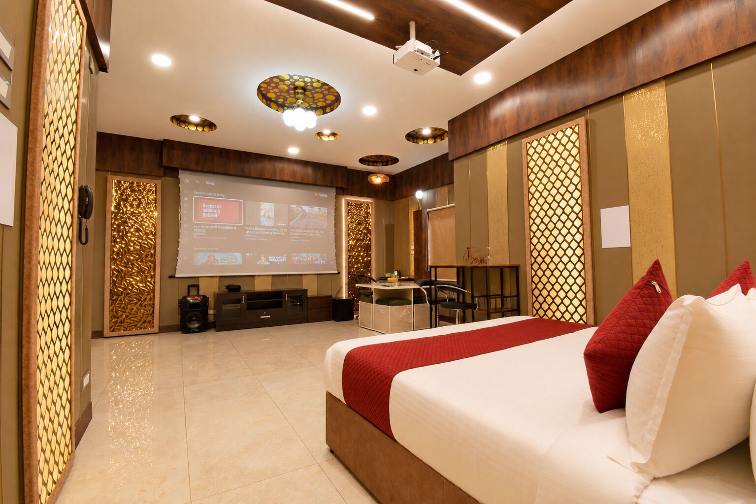 A hotel room with a large bed, a desk with a chair, a projector screen, and a decorative wall panel.