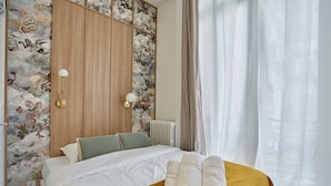 A neatly made bed with rolled towels, a wooden wardrobe, and a floral-patterned wall.