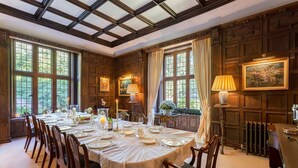 A formal dining room with a long table set for a meal, wooden paneled walls, and large windows with curtains.
