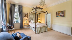 A four-poster bed in a room with a large window, a chair, and a painting on the wall.