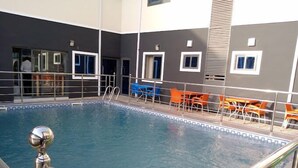Una piscina interior con barandilla metálica, rodeada de sillas anaranjadas y azules, y un edificio de paredes grises y blancas.