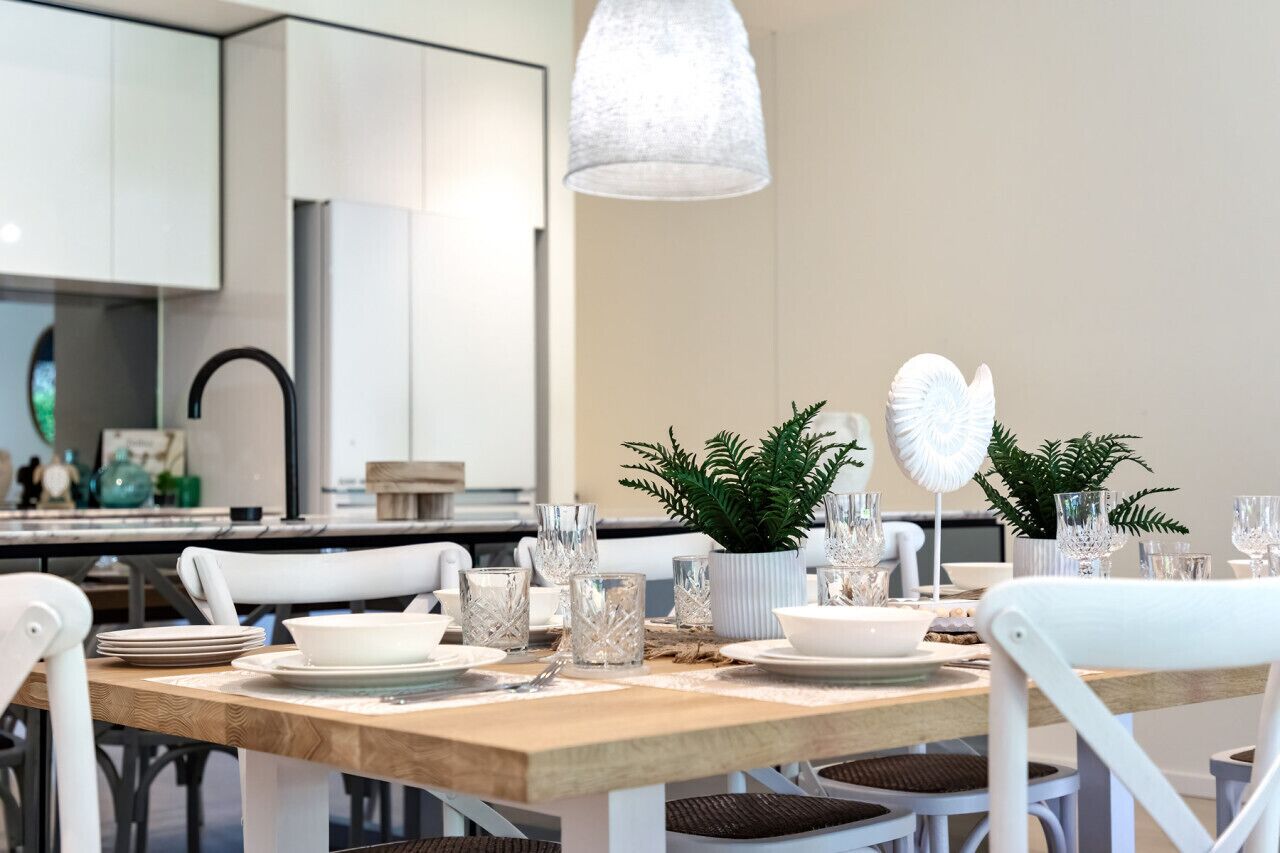 Uma área de jantar moderna com uma mesa de madeira posta para dois, cadeiras brancas e uma luminária pendente.
