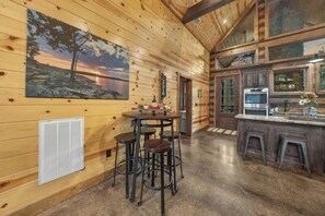 A wooden cabin interior with a kitchen area, a dining table with stools, and a painting of a sunset. - Private Resort Retreat: Pool, Sauna, Golf + Pond (Broken Bow)
