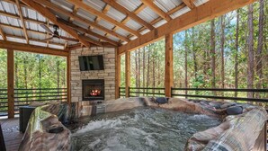 Un jacuzzi con chimenea de piedra, rodeado por una terraza de madera y un bosque al fondo.