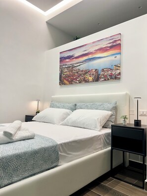 Une chambre moderne avec un grand lit, du linge de lit blanc, des coussins à motifs, une table de chevet avec une lampe et un tableau représentant un paysage urbain accroché au mur.