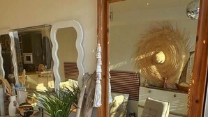 Une pièce avec une commode blanche, un grand miroir, une plante suspendue et un panier en osier contenant des bûches de bois.