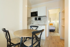 Cocina compacta con microondas, cafetera y lavabo. Hay un comedor con una mesa redonda y sillas. Se ve una cama en la habitación contigua.